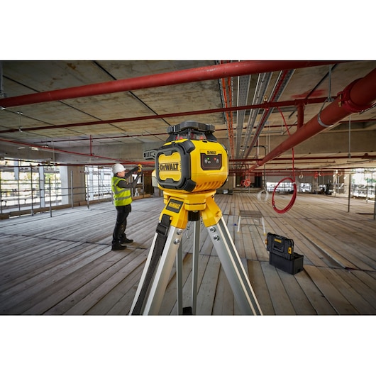 DEWALT Alkaline Rotary Red Beam Laser on tripod on worksite, worker is setting up detector in background.