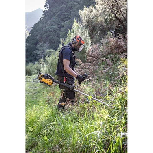 Worker with safety helmet with face visor, ear defenders and gloves using the DEWALT 54V Brush Cutter(Bull Handle) with deluxe harness to cut through dense grass area.