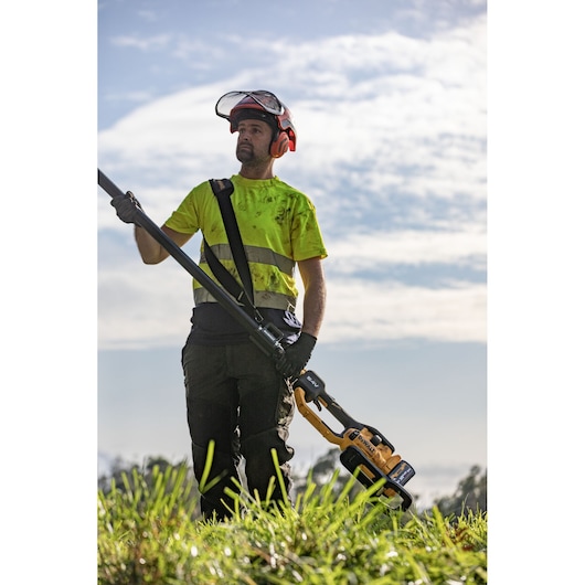 Worker in high visibility top and helmet with visor up and safety harness getting ready to use the extended DEWALT 54V Tele Pole Saw 4.0M 30CM on tree in open large field.
